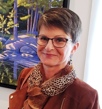 Image of Susan Hay, wearing dark rimmed glasses, short brown hair and a cheetah patterned scarf. Standing in front of her artwork.